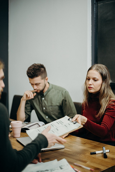 People around a desk, a woman holding showing a clipboard with information on.